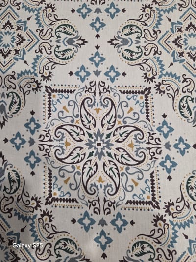 This image presents a detailed close-up of a patterned textile, described as being located in the city of Stroevo, Bulgaria. The fabric exhibits a complex, repeating design on an off-white or cream background, consistent with a woven material. The pattern draws from folk art and traditional paisley motifs, featuring a central quatrefoil-like arrangement of four dark brown paisley shapes with dark green inner accents, surrounded by small gold and blue dots. Surrounding this central motif are various decorative elements, including elegant swirling tendrils in dark brown and light blue, dotted lines in gold and dark brown forming borders, and geometric elements like blue zigzag patterns and small four-petal blue shapes. The color palette primarily consists of dark brown, several shades of blue (light, teal, and dusty gray-blue), dark green, and gold or ochre accents. The textile displays a visible texture, indicating a fabric like cotton or canvas. The image is evenly lit, and a "Galaxy S25" watermark is present in the lower-left corner.