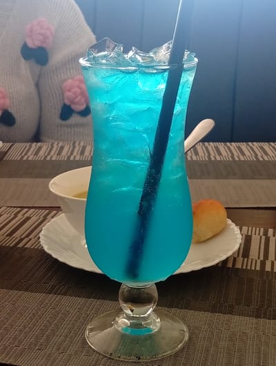 The scene is set indoors at an establishment in Murang'a, Kenya, featuring a table laid with a meal. A prominent, clear glass hurricane-style goblet filled with a vibrant blue, icy beverage and a black straw occupies the foreground. Condensation beads are visible on the exterior of the glass. This goblet rests on a dark grey woven placemat with a subtle striped pattern. Directly behind the drink, on a white plate with a fluted edge, is a small, golden-brown bread roll. To the left of the bread roll, a white bowl containing a pale yellow liquid, likely soup, is partially visible, with a silver spoon resting on its edge. In the blurred background to the left, a person is partially visible, wearing a light-colored, possibly off-white, knitted garment adorned with pink, flower-like appliques that have green accents. The overall background consists of dark, likely wooden or upholstered panels. A bright light source is present from the upper right, creating a slight glare.