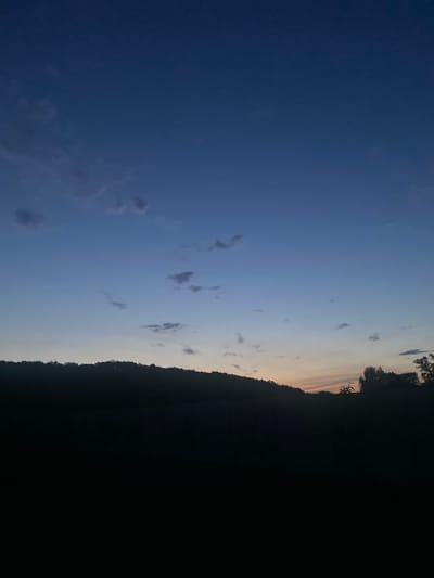 The scene depicts an outdoor landscape, observed during twilight or dawn, in the presumed location of Petrivka, Ukraine. The sky occupies approximately the upper two-thirds of the vertical frame, displaying a color gradient that transitions from a dark blue at the zenith to a lighter blue in the middle sky, culminating in a narrow band of faint orange and yellow light along the horizon. Several small, dark gray clouds are scattered across the blue portions of the sky, primarily in the upper left and central areas. A dark, undulating silhouette, consistent with a distant treeline or hilly terrain, forms the horizon line. The lower one-third of the image is uniformly dark, obscuring any specific details of foreground features or ground cover. No human figures, animals, man-made objects, or specific actions or interactions are discernible within the frame.