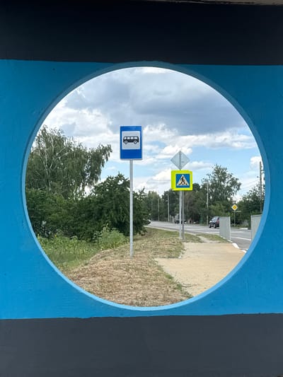 The image, framed by a circular opening in a blue and black structure, captures a view of a street scene in Shulhynka, Ukraine. On the right side of the view, a paved sidewalk runs parallel to a road. Across the road, a grassy area and trees are visible. A black car is parked on the right side of the road. On the left side of the road, a bus stop sign and a pedestrian crossing sign are prominently displayed. The sky above is filled with clouds. The foreground is occupied by the blue and black painted structure, which forms the circular frame. The overall impression is that of a sunny day with a clear view of urban infrastructure and surrounding greenery.