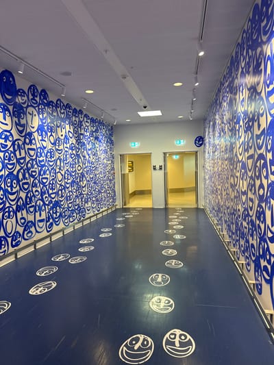 The image depicts a brightly lit, modern interior hallway with a distinct artistic theme. The hallway features a dark blue floor and a white ceiling, both heavily adorned with blue and white graffiti-style artwork. The ceiling is covered in a repeating pattern of abstract blue shapes that resemble stylized faces or characters. The floor on the left side of the hallway is also covered in a dense arrangement of these blue characters.
On the right side of the hallway, the dark blue wall is decorated with a repeating pattern of white circular decals, each featuring a single stylized face with distinct eyes and a smile. These white faces are spaced out, creating a sense of movement or progression down the hall.
In the center of the hallway, there are two sets of glass doors leading into a brightly lit, yellow-toned interior space, likely an elevator lobby or waiting area. Above the doors, green "exit" signs with directional arrows are visible.
The lighting in the hallway appears to be artificial, with track lighting visible along the left wall and recessed circular lights. The overall impression is that of a contemporary public space that has been transformed by vibrant street art. No people are visible in the image. The location is identified as Kastrup, Denmark.