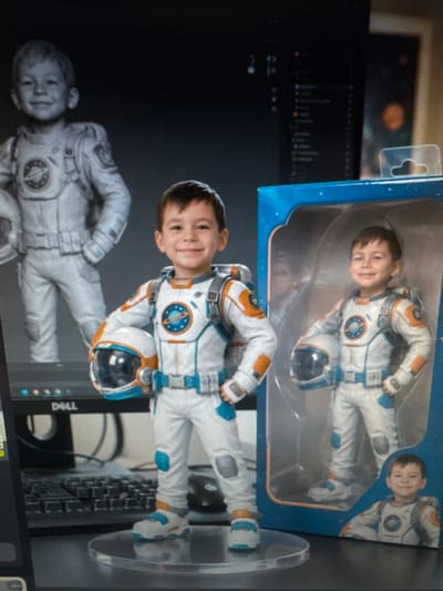 The image displays a still life arrangement on a desk. In the foreground, a boy dressed in a white and orange astronaut suit stands on a clear plastic base. He is holding a white and blue astronaut helmet in his left hand. To the right of the boy's figure is a toy package, also featuring a boy in an astronaut suit. In the background, a computer monitor with a dark screen displays lines of code, and a black keyboard is visible. A black computer mouse rests on the desk. The overall scene is indoors, with a dark backdrop. The location is the city of Protasovo, Russia.