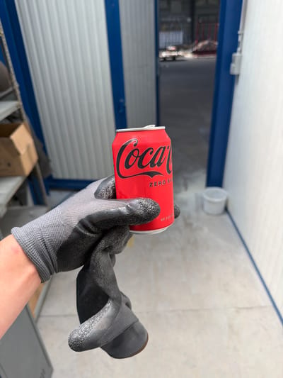 A gloved hand holds a red Coca-Cola Zero Sugar can. The can has the iconic Coca-Cola logo in black script and reads "ZERO SUGAR" below it. The hand is wearing a grey work glove with black textured grip on the fingertips and palm. The background is a light-colored, industrial-looking space with corrugated metal walls and blue trim. In the distance, a large open doorway reveals a bright, blurred outdoor area with what appears to be construction or industrial equipment. A small white bucket is visible on the floor in the middle ground to the right. The overall scene suggests an industrial or storage area, possibly within the city of Mol, Belgium, given the context.