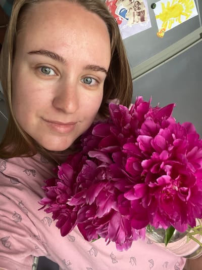 In a medium close-up, a young woman with light brown hair and blue eyes smiles at the camera while holding a large bouquet of vibrant pink peonies. She is wearing a light pink shirt adorned with a repeating pattern of stylized unicorns. The background appears to be a refrigerator door, with a few children's drawings and stickers visible. One drawing depicts a yellow sun-like creature with four legs and what appears to be a red body. Another appears to be a purple sheet with various collage elements, including a blue sticker with Russian text that reads "ДРАТУТИ". The overall lighting is soft, suggesting an indoor setting, possibly in a home in Chaikovsky, Russia.