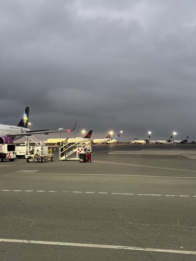 The image shows an airport tarmac, likely at night or in low light, with several aircraft lined up. Various ground support vehicles are parked near the aircraft, including passenger boarding stairs and service vehicles. The tails of the planes are visible, showing livery details of Volaris, Air Canada, and other airlines. The tarmac is marked with white lines. The location is likely in or near Apodaca, Mexico, given the presence of the Volaris aircraft. The sky is overcast.