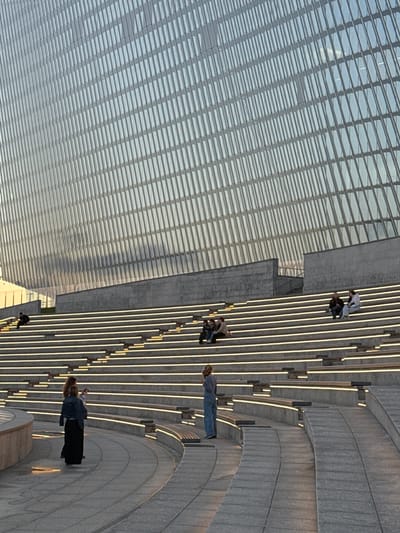 In Alyumino, Russia, an outdoor amphitheater with tiered seating is illuminated by embedded lights. Several people are visible: one person stands near the bottom left of the seating area, facing away from the camera and looking towards the stairs. Further up the stairs, another person stands facing the camera, holding a phone. On the higher tiers of seating, small groups of people are seated and conversing. The background features a modern building with a glass facade, reflecting the sky and surrounding environment. The overall scene depicts a public space in the evening, with people enjoying the ambiance.