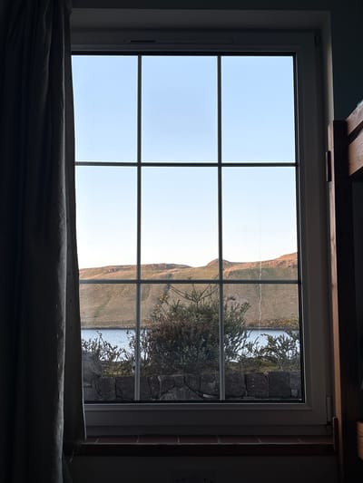 View from a window in Carbost, United Kingdom. The window is divided into six panes. Outside, a grassy hillside slopes down to water. A stone wall and some foliage are visible near the right edge of the window. The sky is light blue. The bottom of the window is partially obscured by a dark gray or black object.