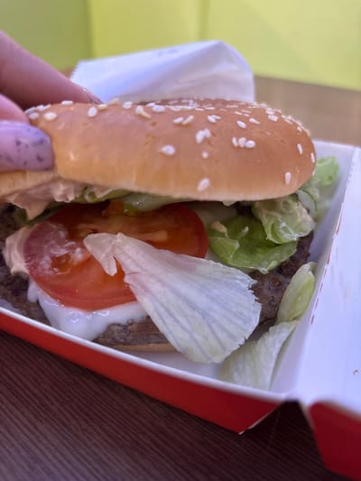 A close-up, eye-level shot shows a person's hand with speckled purple nail polish reaching towards a hamburger. The hamburger is partially enclosed in a white paper wrapper within a red and white cardboard box. The burger has a sesame seed bun, lettuce, a thick slice of tomato, and a meat patty. The background is a blurry, bright green wall and a wooden surface. The image is likely taken indoors, potentially at a restaurant or fast-food establishment in Shendzhii, Russia.