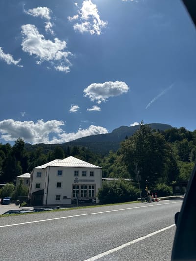 In Berchtesgaden, Germany, a two-story building with a white facade and a light-colored roof stands next to a road. The building has numerous windows and a sign that reads "SALZBERGWERK". Behind the building, a dense forest covers a mountainside, which transitions into a higher, more rugged mountain peak in the background. The sky is blue with scattered white clouds, and the sun is visible, casting light on the scene. The road in the foreground has white lane markings and appears to be a well-maintained asphalt surface. Traffic cones and a traffic signal are visible on the side of the road. Several cars are parked near the building.