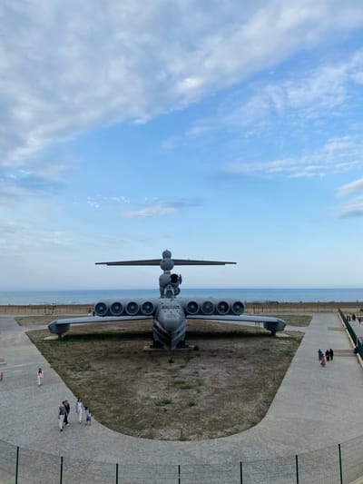An aerial view captures a large, grey ekranoplan, an ekranoplan with eight engines mounted on its wings, displayed on a patch of dry grass near a paved walkway. The ekranoplan is positioned facing forward, with its large wings spread. Behind the ekranoplan, a calm, blue ocean stretches to the horizon, meeting a pale blue sky with scattered white clouds. Several people are walking on the paved pathway in the foreground, observing the ekranoplan. The environment appears to be a public space, possibly a park or museum, with a fence in the lower foreground. The location is identified as Arablyar, Russia.