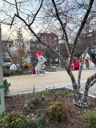 A public park features decorative light sculptures including a unicorn, a rabbit, and some gift boxes on a sandy pathway. Bare tree branches, some wrapped with string lights, frame the scene. A brick building stands in the background under a partly cloudy sky. Green foliage lines the path's edge. Decorative light orbs hang on lower branches.