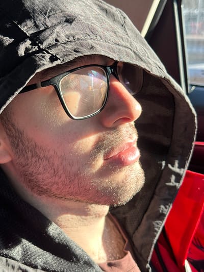 A male individual is depicted in a close-up profile shot, situated inside what appears to be a vehicle. He is positioned towards the left of the frame, facing right, with strong sunlight illuminating the right side of his face and neck. He wears dark-framed glasses, the lenses of which display prominent glare, streaks, and iridescent chromatic reflections. A short, light-colored beard or stubble covers his jawline and upper lip area. His lips are full and slightly parted. He is wearing a dark-colored, textured hooded garment, with the hood pulled up to frame his head. Beneath this, a reddish-brown or mauve-colored garment is visible around his neck. The immediate background to the right shows a blurred red surface, likely an interior car component, with a glimpse of an exterior window further right, through which indistinct bright shapes and light are visible. The scene occurs during daytime in the city of Мещерино, Russia.