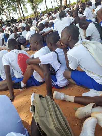 A high-angle, medium shot captures a gathering of young men, likely cadets, dressed in white t-shirts and blue shorts, seated on reddish-brown dirt ground under a canopy of sparse trees. The foreground is dominated by their legs and feet, clad in white socks and various athletic shoes. In the center, a cadet with a red drawstring bag slung over his shoulder is visible, his right arm resting on his thigh. To his right, another cadet, also in white and blue, is partially obscured, with his arms resting on his knees. The background reveals a larger group of similarly dressed individuals seated in rows, their faces turned in various directions, suggesting an organized assembly. A military vehicle is visible in the far distance, along with a few scattered buildings and more trees, hinting at an outdoor training or assembly ground, potentially within or near the city of Barakin Rafin Gora, Nigeria. The overall atmosphere appears calm and orderly, with the sunlight casting shadows on the ground.