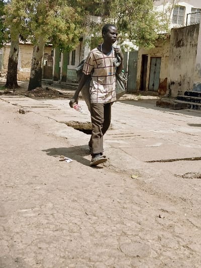 The image captures a young individual walking on a cracked, unpaved and partially paved thoroughfare in front of Kungulu School, Zambia, under direct sunlight. The individual, appearing male, has dark skin and short dark hair, wearing a light-colored, checkered short-sleeved shirt with a visible Nike swoosh logo, dark trousers, and light-colored open-toed sandals. A white string or necklace is visible around his neck. He carries a large, bulky grey sack over his left shoulder and holds a clear plastic water bottle with a red cap in his right hand. His head is turned slightly to his right. The ground features numerous cracks in the concrete sections and exposed dirt. In the background to the left, two trees with light-colored trunks and green foliage partially obscure a light-colored building, where a sign indicating "KUN" (consistent with Kungulu School) is visible. To the right, additional weathered, light-colored buildings are present, one featuring a dark blue door and another with exterior concrete stairs leading upwards. Strong shadows are cast on the ground, indicating midday or early afternoon conditions.