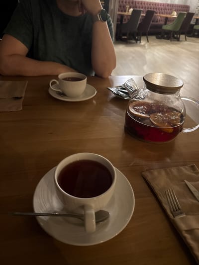 The image shows a person seated at a wooden table in what appears to be a restaurant or cafe. The person is wearing a dark green shirt and a watch. On the table, there is a glass teapot filled with a red beverage containing fruit slices, two white teacups with saucers and spoons, and a tan napkin with cutlery. The location is unspecified, and there is no verifiable connection to Uryupinsk, Russia. The focus is on the beverages and table setting.