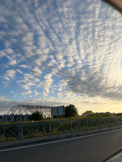 The image depicts a scenic view of the sky with a road and some industrial structures. The sky is filled with fluffy white clouds, stretched across a clear blue expanse. The sun appears to be setting, casting a warm golden glow on the clouds, creating a serene and peaceful atmosphere.
On the right side of the image, a paved road runs parallel to a metal fence. Beyond the fence, there is a lush green grassy area with scattered trees and some buildings in the distance. The architectural style of the buildings suggests a suburban or rural setting.
In the foreground, a large, intricate metal structure is visible, possibly part of an industrial facility or a bridge under construction. Its dark, geometric form contrasts with the soft, organic shapes of the clouds and vegetation. The presence of this structure hints at human activity and development in the otherwise natural landscape.
The overall mood of the image is one of tranquility and natural beauty, enhanced by the warm light of the setting sun. The location is identified as Kholmogorovka, Russia, which adds a geographical context to the scene. There is no visible text in the image.