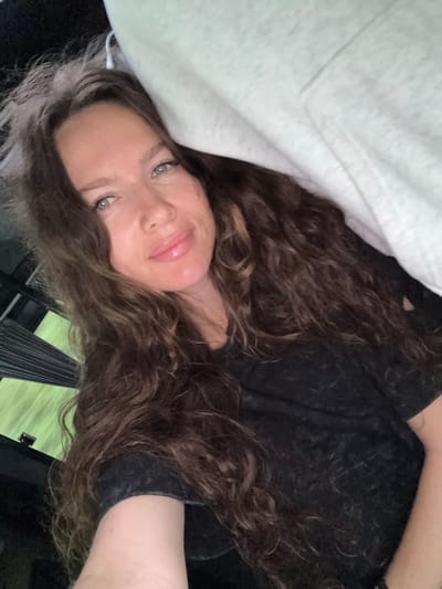 A medium close-up selfie shows a woman with long, wavy brown hair, light blue eyes, and light skin. She is looking at the camera and smiling slightly. She is wearing a dark t-shirt. A light-colored blanket or sheet is draped over her head and shoulder. The background appears to be inside a vehicle, with dark tinted windows showing a blurred green outdoor scene. The image quality is slightly grainy. The selfie was taken in the city of Turbaza Katun', Russia.