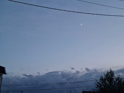The image shows a twilight sky over Novostroyka, Russia. A thin crescent moon is visible in the upper right quadrant of the frame. Several power lines stretch across the sky from left to right. In the lower portion of the image, a dark, overcast cloud bank runs horizontally, with a sliver of a building roof visible on the far left and a dense tree on the far right, also dark against the sky. The overall impression is a calm, late evening scene.