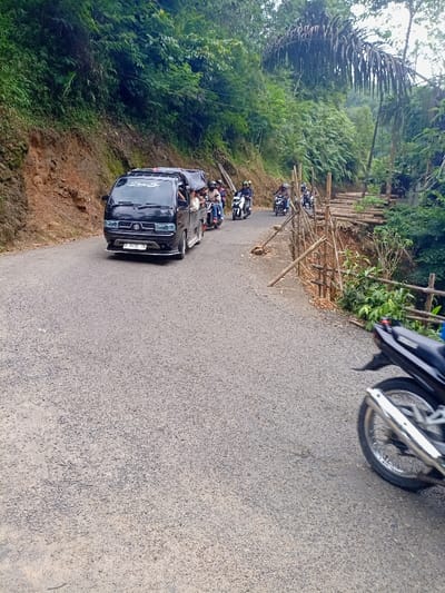A black van is driving on a paved road in Cisewu, Indonesia (latitude: -7.41, longitude: 107.542). There are several motorcycles parked on the side of the road, and a motorcycle is also parked on the right side of the image, partially out of frame. Green vegetation is on both sides of the road. The road is bordered by a bamboo fence on the right, along with a steep embankment. Several people are standing by the van, including two men sitting in the back of the van, facing the driver's seat. The van is facing forward and the driver is seated behind the wheel.  The van has a white "Oney" sticker on the windshield.  There is also a white sticker on the rear window with the word "Suzuk". The image is taken from a street level perspective.
