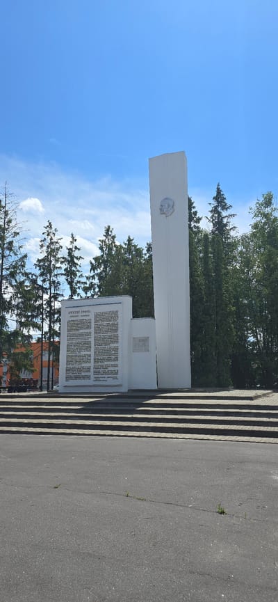 The image shows a war memorial located in the city of Bryansk, Russia. The memorial consists of a tall, white, obelisk-shaped monument with a relief of a man's head on its upper section. To the left of the obelisk is a shorter, white rectangular monument with text inscribed on it, and to the right of that is a smaller plaque. The monuments are situated on a paved area, with several steps leading up to them. Behind the monuments, there are trees and a clear blue sky with some clouds. The foreground shows a section of asphalt road. The overall scene is a public monument area on a clear day.