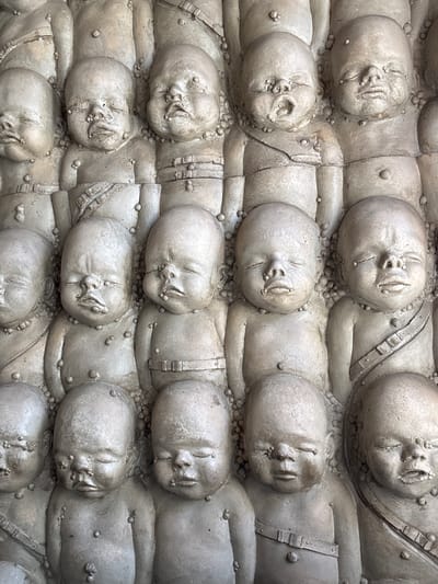 The image depicts a close-up of a sculptural wall composed of numerous figures lying down, each appearing to be a newborn infant. The figures are made of a light gray material, possibly concrete or stone. The figures are arranged in rows and columns, tightly packed together. They appear to be wearing some kind of body armor or straps. There are many small stones or beads that surround the infants. This sculpture is located in Epagny, Switzerland.