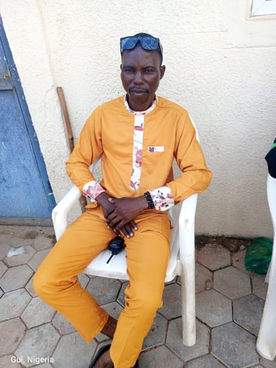 A man is seated in a white plastic chair on a hexagonal-tiled outdoor surface. He is wearing an orange tunic with a floral patterned collar and cuffs, and orange trousers. A pair of black sunglasses is perched on his head. His hands are clasped in his lap, and a black walkie-talkie rests on his trousers. The background consists of a textured, off-white wall and a blue metal door to the left. The scene is identified as being in Kuje, Nigeria.