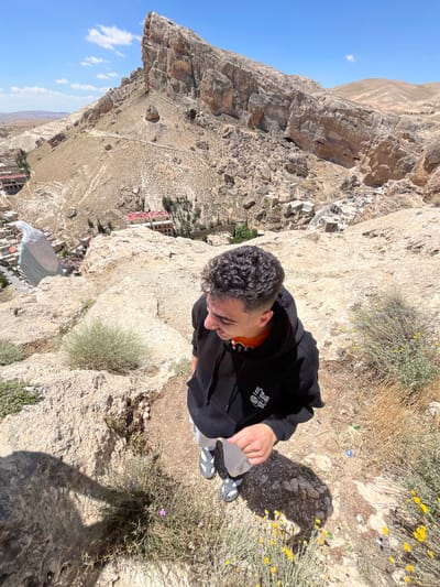 A young man stands on a rocky edge overlooking the city of Ma'loula, Syria. He wears a black hooded sweatshirt over an orange shirt and holds a plastic bag in his right hand. He has short, dark, curly hair. The background shows a steep, rocky landscape and buildings below. The sky is visible in the upper left corner and is blue. The ground near the man has low-lying vegetation.
