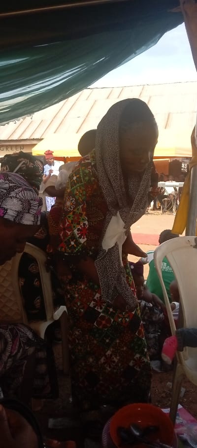 The scene depicts an outdoor gathering occurring under a dark green fabric canopy in Kasuwan Magani, Nigeria. In the foreground, an orange plastic bowl containing multiple metal spoons rests on a purple plastic plate. The central figure is a woman, wearing a multi-patterned long dress with brown, green, red, and white geometric designs, and a dark grey, speckled head covering draped over her head and shoulders. She carries a child on her back, secured by a fabric wrap, with the child's head partially visible. The woman is oriented downwards. To her left, a partially visible individual is seated on a white plastic chair, wearing a dark, patterned head wrap. In the mid-ground, a person wearing a red head covering and a light blue patterned dress stands, holding what appears to be papers, addressing a group of individuals who are partially visible. Further back, an electronic keyboard on a stand is discernible. The background features corrugated metal roofs of structures under a clear sky, and a partial sign displaying the word "GREEN." The ground beneath the canopy consists of compacted earth.