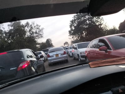 The image captures a scene of heavy traffic from the perspective of someone sitting inside a car, looking out through the windshield. The upper portion of the image is obscured by the interior roof lining and sun visor of the car, and the bottom by the dashboard, which has a brown cloth placed on it.

A long line of stationary or very slowly moving vehicles stretches into the distance. In the immediate foreground on the left, the rear of a dark-colored (possibly black or dark grey) hatchback car is visible, showing its red taillights. Its model appears to be a Mazda Demio, though the text is partially obscured. On the right, the front-right quarter of a red sedan or hatchback is prominent, showing its side mirror with an integrated turn signal.

Further down the line, a white minivan or utility vehicle (possibly a Toyota Probox or similar) is visible behind the red car. Behind it and slightly to the left, another light-colored (possibly silver or light grey) sedan is seen, with its rear facing the viewer. This car has a yellow license plate that appears to read "KBC 901D" or similar, with the "901D" being clearer. Other cars, mostly light-colored, are packed closely together, extending far into the background, where they become less distinct due to distance and slight haze.

The road itself is not clearly visible due to the density of the cars. On either side of the road and in the distant background, numerous trees with dense foliage are visible, suggesting a somewhat suburban or natural setting bordering the road. The sky above is a light, somewhat hazy grey, indicating either early morning, late afternoon, or an overcast day. The overall impression is one of a typical traffic jam during peak hours.
