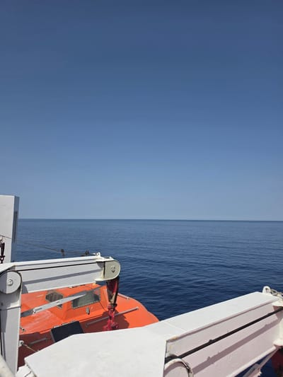 The image shows a view from a vessel at sea, likely a ferry or a cruise ship, with the calm, blue sea stretching to the horizon under a clear, light blue sky. In the foreground, part of the ship's deck is visible, including white railings and equipment. A bright orange lifeboat, suspended from a davit, is prominently featured on the left side of the frame. The lifeboat has two small windows and a black hatch on its top surface. A thick rope is secured to the railing. The scene suggests a bright, sunny day at sea, possibly in the Mediterranean, given the mention of Sol de Mallorca.