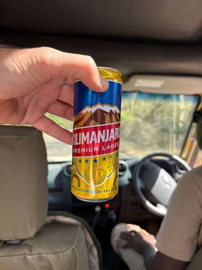 The image displays the interior of a right-hand drive vehicle, located in Lerai Ranger Post, Tanzania. In the foreground, a light-skinned hand holds a slender, blue and gold aluminum can of "Kilimanjaro Premium Lager," which prominently features a mountain graphic and states "4.5% ALC/VOL." The can is positioned in front of a tan upholstered vehicle headrest. In the background, partially visible in the driver's seat, is an individual wearing light-colored clothing; their arm and leg are discernible. The vehicle's dashboard, steering wheel, and a dark roof lining are also visible. Through the front windshield, a blurred sunlit outdoor environment with green foliage is depicted.