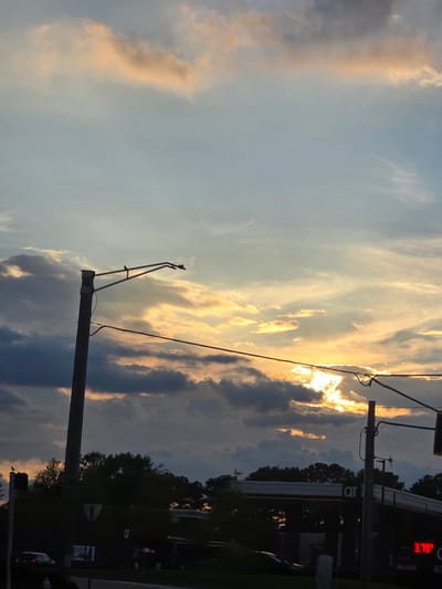 This image captures a sunset over a QT gas station in Norcross, Georgia, United States. The sky is filled with dramatic clouds, with patches of orange and yellow light from the setting sun breaking through. A utility pole with a streetlamp stands prominently in the foreground, silhouetted against the colorful sky. In the background, the QT gas station canopy is visible, along with a sign displaying the price of gas, "3.79". Trees line the horizon, and a few cars are parked or driving on the road in front of the station. The overall scene depicts a common urban landscape at dusk.