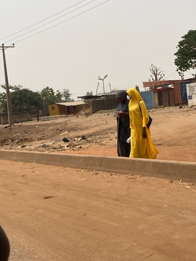 The image captures an outdoor scene in Lapai, Nigeria, under a hazy sky, featuring a reddish-brown dirt road in the foreground. A concrete curb separates the road from an elevated dirt embankment strewn with debris, including dark trash bags.
Two adult females are positioned on this embankment, walking or standing. The individual on the left wears a dark gray, full-length garment and a dark head covering; she is holding a white rectangular object, possibly paper, and appears to be writing on it with a dark pen. The individual on the right wears a bright yellow, full-length garment with a matching head covering, carries a dark bag over her left shoulder, and holds a rolled-up white document in her right hand.
In the background, from left to right, are a wooden utility pole with overhead wires, a dense green tree, a concrete wall, a small, indistinct figure near the wall, a yellow metal container with a sloped roof, and various other structures including parts of buildings, a street light with a solar panel, a blue gate, and a reddish-brown metal container. Sparse vegetation, including a gnarled tree, is also visible.