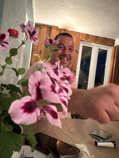 The image depicts an indoor scene, possibly a room or a covered porch. In the foreground, a plant with pink and dark purple flowers is prominently featured, with one large flower obscuring part of the subject. To the left of the plant is a clear plastic bottle with a red cap. The subject, an adult male with short hair, is smiling and looking towards the camera, with his right arm and hand extended into the frame, appearing to be reaching out. The background shows wooden paneling and a window with white frames, suggesting an outdoor view or a room with a rustic aesthetic. On the wooden table in the foreground, there are several objects including a smartphone, a lighter, and a small stack of playing cards. The overall lighting suggests it might be evening or indoors with artificial light. The context of Heinebach, Germany, does not provide specific visual cues within the image itself, therefore, it cannot be incorporated into the forensic description of the scene.