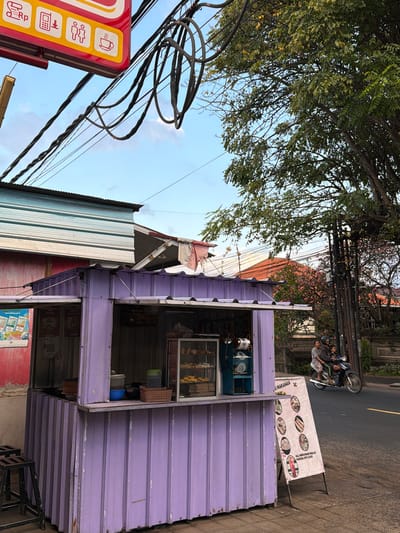 In Kubutambahan, Indonesia, a small, purple food stall stands on a sidewalk next to a street. The stall has a corrugated metal exterior and is topped with a matching roof. Inside, food is displayed in a glass case, and various items are arranged on the counter, including containers and a basket. A sign with pictures of food items and text in Indonesian stands in front of the stall. To the right of the stall, two people on a motorcycle, a man and a child, ride past on the asphalt street. Overhead, a tangle of electrical wires crisscrosses the sky, with a large tree casting shadows. In the background, houses with red tile roofs and other structures are visible. The sky is partly cloudy.