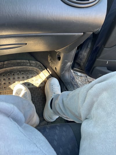 The image presents a ground-level view from within a vehicle, specifically from the passenger side, looking down towards the floor and out an open door. The primary subject is a person's lower legs and feet. The individual is wearing light grey, possibly fleece or textured fabric, trousers and clean white athletic shoes with white laces. Black socks are visible at the ankles. The feet rest on a black rubber floor mat featuring a grid of raised circular patterns. In front of the feet, the lower part of a dark grey dashboard or glove compartment is visible, showing some minor scuffing. To the right, the passenger door is open, revealing a dark blue door panel and the rough exterior ground, which appears to be a dirt or gravel surface with sparse vegetation or debris. Bright sunlight illuminates parts of the car interior and casts shadows, particularly on the floor mat. A section of a dark grey car seat with a subtle diamond pattern is visible in the lower right quadrant. The scene is located in Venets, Bulgaria.