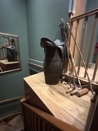 Within an indoor setting in the Borough of Runnymede, United Kingdom, a static decorative display is presented. A dark-stained wooden barrel, secured with two metal bands, stands vertically on a light-toned wooden landing. A dark brown leather saddle rests atop the barrel. Six wooden polo mallets with wrapped handles are leaning against a light wooden balustrade and a dark teal-green wall behind the barrel; three mallets have their cylindrical heads resting on the wooden landing. To the left, a rectangular mirror with an ornate gold-colored frame is mounted on the dark teal-green wall, reflecting a portion of the barrel, saddle, and a descending wooden staircase with dark banisters. Light-colored vertical wooden paneling is visible behind the balustrade, along with the top of a red fire extinguisher in the background. No individuals are present, and no actions are depicted.