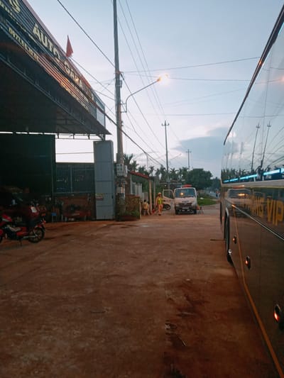 The image shows a street scene in Xuân Long, Vietnam. On the left is a building with a sign reading "AUTO GARA," under a metal awning. A red motorcycle is parked outside. Across the street, a white truck with a light bar on top and "CSCT" is parked. Two individuals wearing yellow reflective vests are standing near the truck. A large bus occupies the right side of the frame, with "VIP" visible on its side. Utility poles and power lines are strung overhead. The street appears unpaved and wet. The sky is overcast.