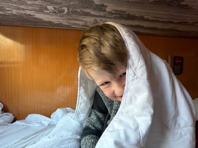 A young, light-haired child with blue eyes is depicted partially concealed beneath a white quilted blanket in an interior setting. The child wears a grey and black striped sweater and peers towards the viewer with a slight smile. The background features light brown wooden paneling on the wall, with white bed linen visible on the lower left. Above, a textured ceiling or upper bunk surface displays a monochrome, cloud-like pattern. To the right, a black vertical object with a red circular prohibition symbol, characteristic of a "no smoking" sign, is mounted on the wall. The scene occurs within a sleeping compartment or bunk area, located in Goričani, Montenegro.