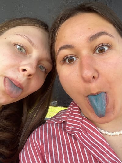 The image shows a close-up, overhead view of two young women. Both have their heads tilted and are sticking out their tongues, which are colored blue. The woman in the foreground has light green eyes and brown hair, and is wearing a red and white striped shirt. The woman behind her has brown eyes and brown hair, and is wearing a white beaded necklace. The lighting suggests it is daytime, and the setting appears to be outdoors, possibly in a park or natural area, given the presence of greenery in the background. The overall mood of the image seems playful and lighthearted. There is no visible text in the image. The location context provided is Рассудово, Russia.