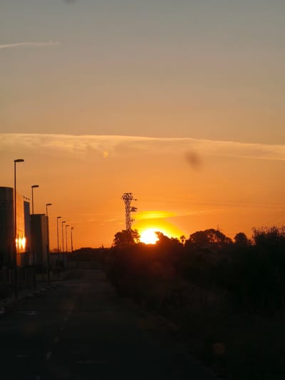 The image depicts a sunset scene on a road in Nàquera, Spain. A straight road stretches into the distance, flanked on the left by modern buildings with vertical metal slats and streetlights. On the right, trees and bushes form a silhouette against the setting sun. An electrical tower stands in the center, partially obscuring the sun. The sky is a gradient of orange hues, brighter near the horizon and fading into a pale blue towards the top. Buildings on the left side reflect the sunset glow.