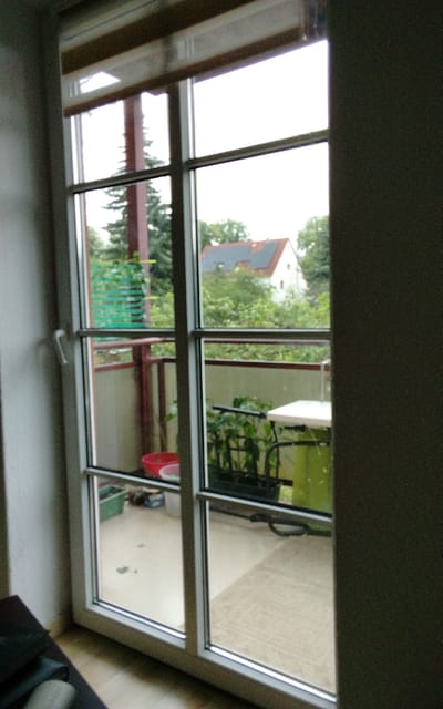 The image shows a view from inside a building, looking out through a large window or glass door. The window is divided into multiple panes by white frames. Outside, the scene depicts a balcony and a view of houses and trees.
On the balcony, there are several objects visible. In the lower left section of the balcony, a red basin is present. Further to the right, there are some green planters containing plants. A small white table or shelf unit is also on the balcony, with a green object, possibly a bin or cover, behind it. The balcony floor appears to be paved and there is some clutter scattered around, including a green rectangular container and some darker items. A railing runs along the front of the balcony.
Beyond the balcony, the view includes houses with red and gray roofs, surrounded by lush green trees. The sky is overcast, suggesting it is daytime, likely morning or afternoon, with dull weather. The overall atmosphere is calm and domestic.
The image is taken from an interior space, with a wall and part of a door frame visible on the left and right sides respectively. The lighting is natural, coming from outside. There is no visible text in the image. The context suggests this is a residential setting in Halberstadt, Germany.