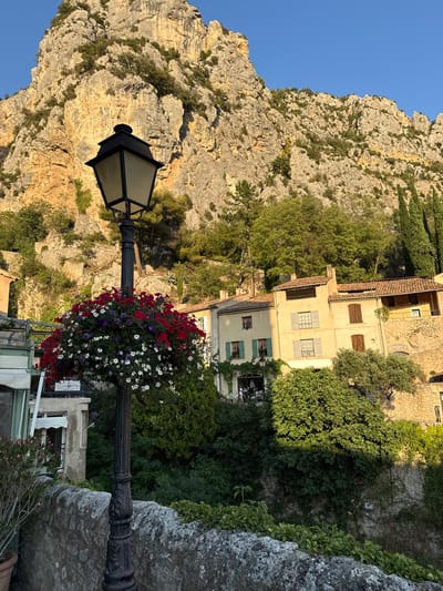 A lamppost decorated with a hanging basket of red, white, and purple flowers stands in front of a rocky mountain and a village in Moustiers-Sainte-Marie, France. The lamppost is black and ornate, with a square lantern at the top. The hanging basket is full of vibrant flowers, creating a beautiful contrast with the natural landscape. Behind the lamppost, the village buildings are made of stone and stucco, with red tile roofs. The mountain is rugged and steep, with trees and vegetation growing on its slopes. The sky is clear and blue, and the sun is shining, casting a warm glow on the scene.
