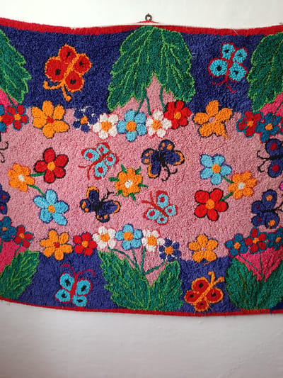 A rug with a colorful floral and butterfly pattern hangs on a white wall in a room. The rug has a red border and is predominantly pink, blue, and green. It features various embroidered flowers in shades of red, orange, blue, and white, as well as numerous butterflies. The scene does not contain any people or demonstrate any actions or interactions. The room appears to be undecorated, with the focus solely on the hanging rug.