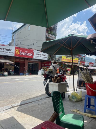 This is a street-level, medium-angle shot of a food stall in Tân Quang Commune, Vietnam. The scene is dominated by the presence of large green umbrellas providing shade over the sidewalk. In the mid-ground, a person in a white t-shirt is leaning over a red motorcycle, which is loaded with what appears to be food items in white containers. The motorcycle has a license plate with the characters "43-AB 973.83".

Various signs are visible on the buildings lining the street, including "BIZ SAIGON" and "HANH THUY COM PHO - COM RANG DU MON". The street itself is paved, with a sidewalk made of concrete tiles. Scattered on the sidewalk are plastic chairs, a red bucket, and a yellow plastic bag. In the background, there are multi-story buildings, some with balconies, and power lines. The sky is blue with fluffy white clouds, indicating daytime. The overall impression is a busy, everyday scene in a Vietnamese urban or semi-urban area.