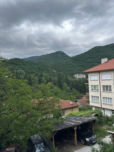 The image captures a residential area in the city of Narechenski Bani, Bulgaria, under a cloudy sky. In the foreground, a weathered, open-sided carport shelters a black car. To the left of the carport, the hood of a dark red car is visible. Behind the carport, the roofs of several houses with red tiles are partially obscured by lush green trees.
Dominating the midground and background are rolling, densely forested hills, characteristic of the surrounding mountainous terrain. The foliage appears verdant and healthy. A multi-story building, likely an apartment complex, is visible nestled amongst the trees in the distance. The overall atmosphere suggests a tranquil, somewhat overcast day in a natural setting. The architecture of the visible buildings points towards a European style. There are no people or animals depicted in the image.