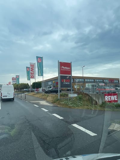 The image depicts a street scene in Lauterbach, Germany, with a focus on a commercial area. On the right side of the road, a large building houses several businesses, including "REWE" and "REWE City," indicated by prominent signage. Adjacent to REWE, there is a "REWE" sign on a concrete barrier, and another "REWE" sign on the building itself. Also visible on the building are signs for "ACTION" and "VIVA FITNESS." A large red sign with white text reads "Philipps," with the subtitle "DER DISCOUNTER FÜR HEIM & GARTEN."

Several flags are planted along the roadside. Three vertical flags with a green background and red circular logo read "BEME." Another flag, also green with a red circular logo, has white text that appears to be the same as the other "BEME" flags but is harder to decipher due to its orientation. Further down the road, a flag with red text on a white background reads "REVV," and another partially obscured flag displays "ACTION."

On the left side of the road, a white panel van is parked, facing away from the camera. A sign on the road indicates "Trocken / Ruckel," possibly denoting a business name or a warning. The road itself is asphalt, with white lane markings. The sky is overcast with a cloudy, grey appearance. The surrounding environment includes trees and vegetation along the roadside. Cars are visible parked in front of the businesses and on the road.