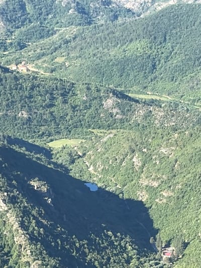 An aerial, high-angle, full shot captures a mountainous, verdant landscape in Kućišta, Montenegro. The image displays steep, forested slopes, with some rocky outcrops visible. A small, blue body of water is nestled in the valley. In the upper left quadrant, a cluster of red-roofed buildings is situated amidst the trees. A winding dirt path is discernible, leading towards the buildings. The scene is bathed in sunlight, casting shadows across the terrain. No people or animals are visible.