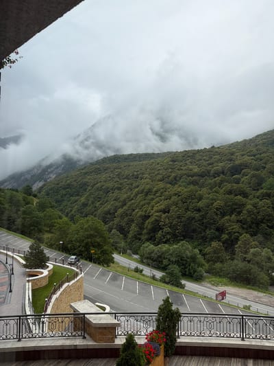 The image captures a mountainous landscape with dense green forests covering the slopes. The sky is overcast with thick clouds obscuring the upper parts of the mountains, creating a misty atmosphere. In the foreground, there is a paved parking lot with white markings, and a road runs alongside it. A silver car is parked in the lot. Adjacent to the parking lot, there is a curved retaining wall made of stone, topped with a decorative metal railing. Beyond the wall, there are well-maintained grassy areas and some planters with red flowers. The scene suggests a viewpoint from a building or balcony with a wooden floor and another ornate railing in the immediate foreground. The overall impression is one of natural beauty with man-made structures integrated into the environment. This location is identified as being in the city of Dzivgis, Russia.