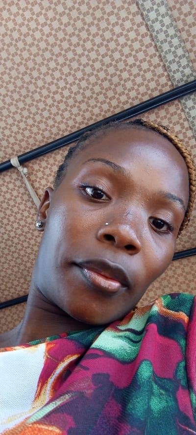 A close-up photograph depicts a woman with dark skin, wearing a floral-patterned garment with red, green, and white hues. She has a nose piercing and small earrings. Her hair is styled in braids with blonde ends. The background reveals a patterned ceiling, likely inside a vehicle, with a black metal frame partially visible. Given the request to place the scene in Uke, Nigeria, the woman is possibly inside a commercial transport vehicle like a "keke" or taxi, which are common modes of transport in many Nigerian cities. The location is inside the city of Uke, Nigeria.